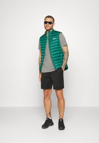 Grön pufferväst över grå t-shirt, svarta shorts och svarta skor. Solglasögon med gröna och lila detaljer. Enkel, funktionell design.