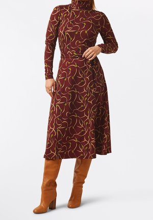 Woman wearing a maroon midi dress with abstract yellow-green pattern, long sleeves, high neck, and a fabric belt, paired with tan knee-high boots.