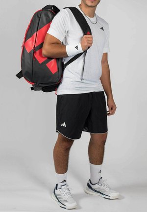 Junger Mann trägt weiße Sportbekleidung und schwarze Shorts, mit einem großen schwarzen und roten Sport-Rucksack über einer Schulter.