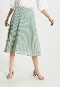 Gonna plissettata verde chiaro con motivo a pois dorati, vita aderente e design morbido. Abbinata a sandali con tacco a blocco beige.