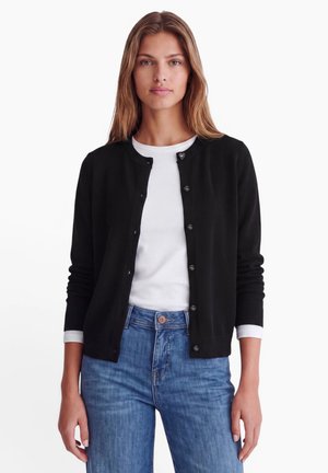 Frau trägt einen schwarzen, geknöpften Cardigan über einem weißen Hemd und blauen Jeans, stehend vor einem schlichten hellen Hintergrund.