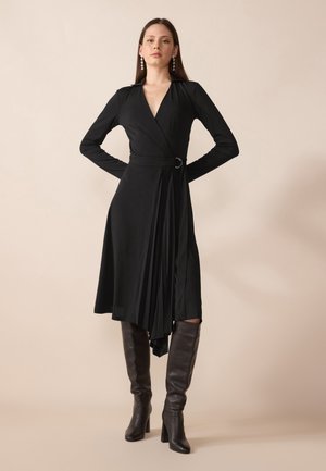 Black wrap dress with long sleeves, a v-neck, pleated front, and a circular belt detail, paired with knee-high brown leather boots.