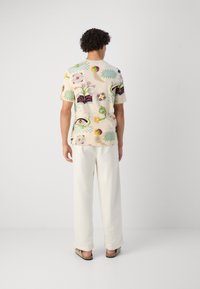 Short-sleeve shirt in cream fabric featuring a floral and abstract print in vibrant colors. Worn with loose-fitting white trousers.