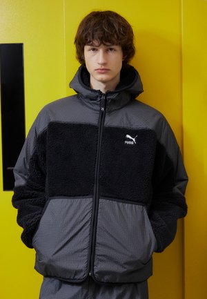 Jachetă neagră și gri cu un superior din polar texturat și o secțiune inferioară în carouri, dotată cu glugă și fermoar frontal. Logo-ul Puma este afisat.