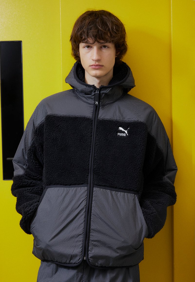 Čierna a šedá bunda s textúrovanou fleecovou hornou časťou a kockovanou dolnou časťou, so kapucňou a predným zipsom. Zobrazené logo Puma.