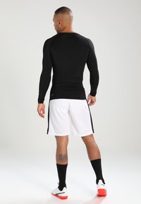 Langærmet sort atletisk skjorte parret med hvide shorts med sorte sidelister. Rund hals, tætsiddende design, glat tekstur og sporty sokker.