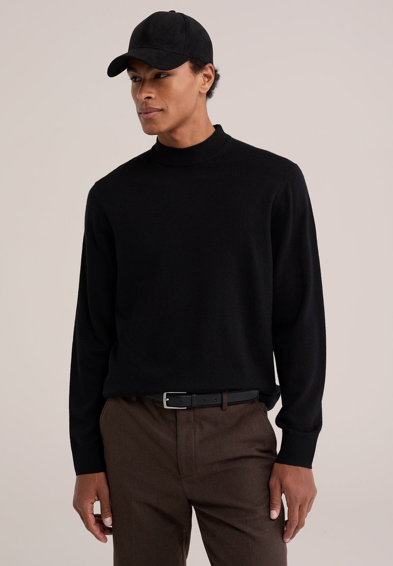 Homme portant une casquette noire, un pull noir à manches longues, un pantalon marron et une ceinture noire, debout devant un fond clair uni.
