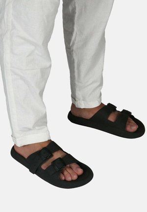Sandales noires avec deux boucles réglables, semelle plate et design ouvert. Portées avec un pantalon en lin blanc, doté d'un ourlet à revers.