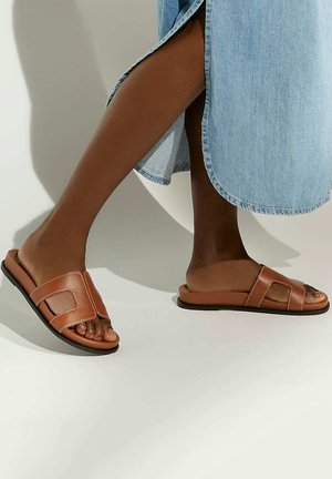 Bruine leren slidersandalen met twee decoratieve banden, een platte zool en witte stiksels, gecombineerd met een lichte denims nederdel.