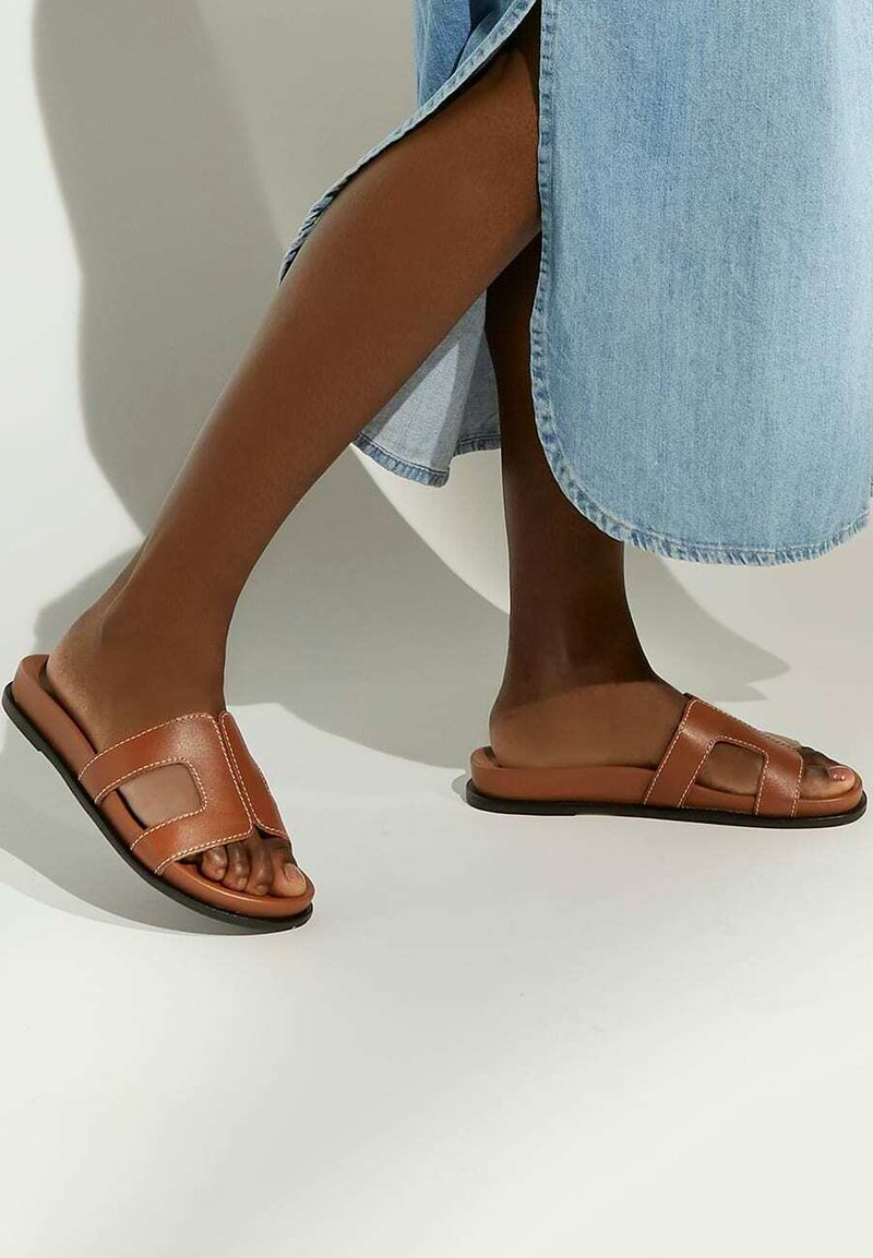 Bruine leren slidersandalen met twee decoratieve banden, een platte zool en witte stiksels, gecombineerd met een lichte denims nederdel.