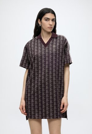 Jonge vrouw met lang donker haar draagt een donker polokleed met korte mouwen en een klein logo op de borst, staand tegen een witte achtergrond.