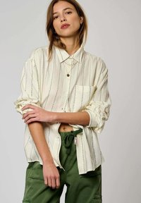 Chemise beige clair à boutons avec des rayures verticales vertes et une poche poitrine, associée à un pantalon vert à cordon de serrage. Finition en tissu doux.