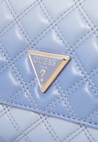 Borsa sintetica trapuntata blu chiaro con un logo triangolare dorato recante la scritta "GUESS" e un punto interrogativo.