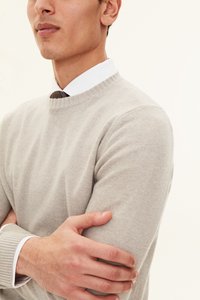Pull en laine gris clair avec un col rond et des poignets côtelés, porté par-dessus une chemise blanche à col et une cravate à motif sombre.