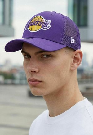 Junger Mann mit lila Los Angeles Lakers-Basecap und weißem Hemd, ernst blickend mit unscharfem urbanem Hintergrund.