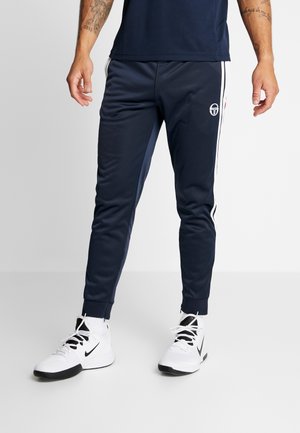 Uomo che indossa pantaloni da ginnastica sportivi blu navy con strisce laterali bianche e sneakers Nike bianche su uno sfondo chiaro uniforme.