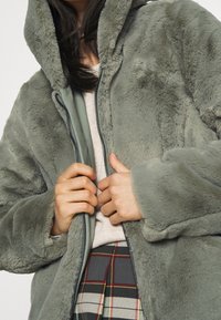 Person som bär en mjuk grå huva av konstpälsjacka, beige topp och rutmönstrad kjol, som fäster dragkedjan på jackan med båda händerna.