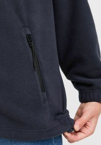 Marineblå fleecegenser med glidelås-lomme på siden, elastiske mansjetter og justerbar hem. Myk tekstur og uformelt design.