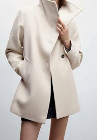 Cappotto in lana beige con colletto alto, due tasche anteriori e chiusura a singolo bottone. Texture liscia e vestibilità aderente, che arriva a metà coscia.