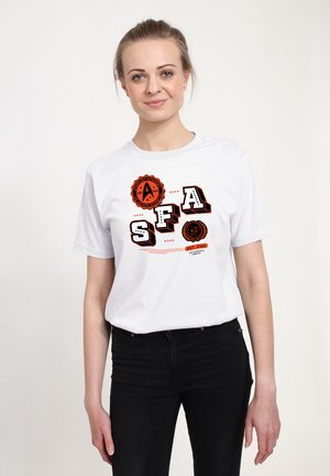 Frau trägt ein weißes T-Shirt mit schwarzen und orangefarbenen "SFA" Buchstaben und weltraumbezogenen Aufnähern, kombiniert mit schwarzen Hosen, steht vor weißem Hintergrund.