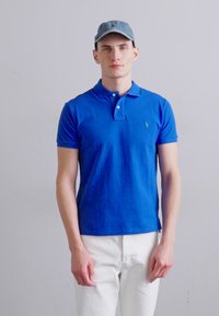 Sinine polosärk puuvillast kangast slim fit lõikega, kolme nööbiga käeauk ja väike tikandiga logo. Kandmiseks valgete püksidega.
