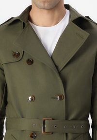 Homme portant un trench-coat olive à double boutonnage avec des boutons marron et une ceinture assortie sur un t-shirt blanc à col rond.