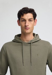 Felpa con cappuccio verde oliva realizzata in tessuto morbido, con cordini regolabili e un piccolo logo ricamato sul petto.