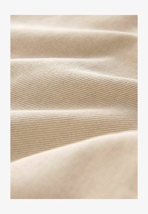 Bēšas krāsas audums ar gludu, mīkstu faktūru un diagonālu ribu rakstu, uz kura redzamas vieglas krokas un locījumi.