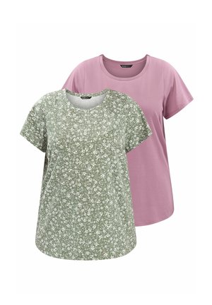 Mujer con cabello rubio que lleva una camisa verde con estampado floral de manga corta, vaqueros azul claro con dobladillo y zapatillas blancas, de pie con una mano en la cadera.