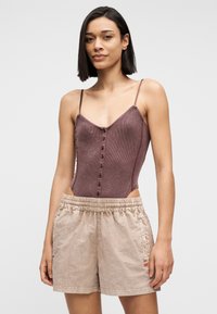 Γυναίκα με ίσια μαλλιά μέχρι τους ώμους, που φοράει καφέ αμάνικη μπλούζα με κουμπιά και μπεζ σορτς με ελαστική μέση, στέκεται μπροστά σε απλό φόντο.