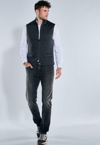 Dunkelgraue, ärmellose Weste mit einem Frontreißverschluss und zwei Taschen. Getragen über einem weißen Langarmshirt und kombiniert mit dunklen Jeans und Turnschuhen.