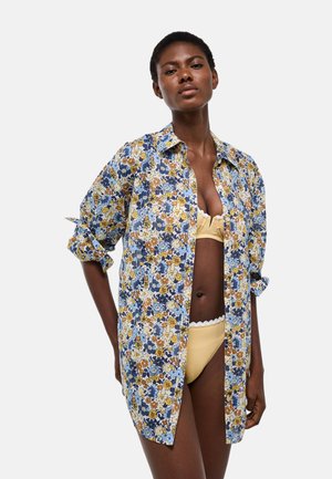 Femme portant un bikini jaune et une chemise à manches longues fleurie bleue, jaune et blanche, debout avec une main dans le dos sur fond blanc.