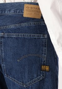 G-Star RAW traperice s teksturiranim plavim završnom obradom. Imaju kožni uzorak na struku i žuti logotip na stražnjem džepu.