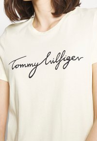 T-shirt en coton crème avec un logo noir en cursive portant l'inscription "Tommy Hilfiger." Manches courtes et design classique à col rond.