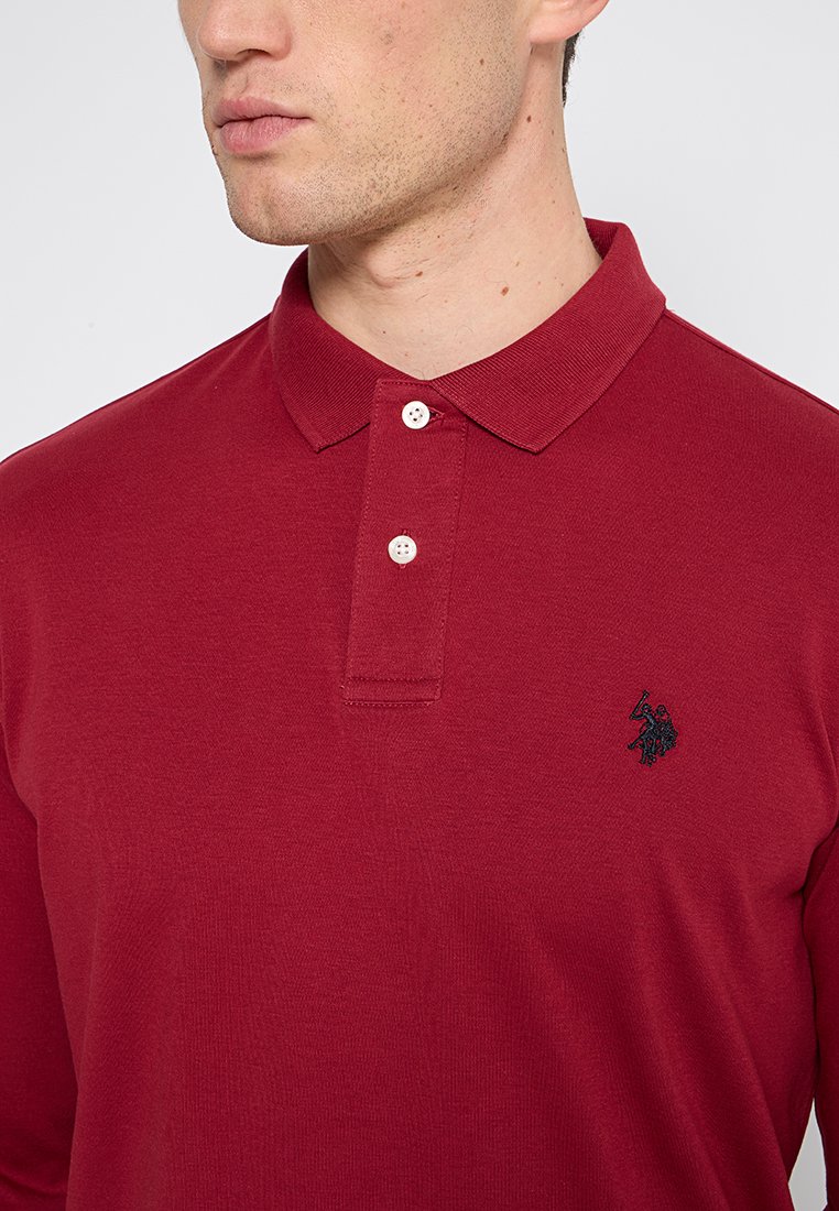 Homme portant un polo à manches longues rouge avec deux boutons blancs et un petit logo brodé foncé sur la poitrine.
