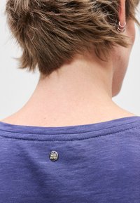 Μωβ μπλουζάκι που φαίνεται από την πλάτη, με στρογγυλή λαιμόκοψη, υφή υφάσματος και μια μικρή μεταλλική λεπτομέρεια λογότυπου κοντά στη λαιμόκοψη.