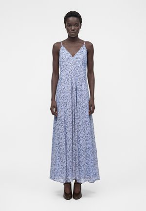 Kobieta stojąca, ubrana w długą, jasnoniebieską sukienkę w kwiaty z cienkimi ramiączkami i czarne, zakryte obcasy, na jednolitym białym tle.