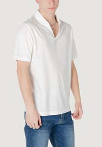 Maglietta polo bianca in cotone con scollatura a V profonda, maniche corte e fit comodo, abbinata a jeans blu, mostrata dalla vita in su.