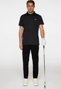 Musta poolopaita, jossa on kaulus ja pieni logo rinnassa. Päällä mustat housut ja valkoiset kengät, pitää kädessään golfmailaa.