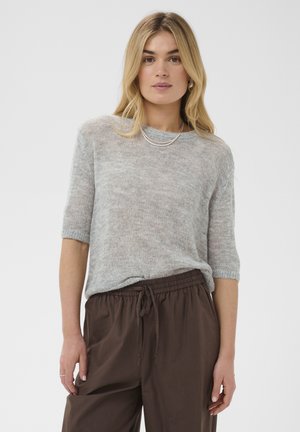 Femme aux longs cheveux blonds portant un pull à manches courtes gris clair et un pantalon marron ample avec cordon, debout devant un fond clair uni.