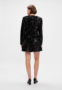 Svart paljettklänning med långa ärmar, med en åtsittande överdel och flödande kjol. Slät textur med reflekterande paljetter, tillsammans med svarta klackar.