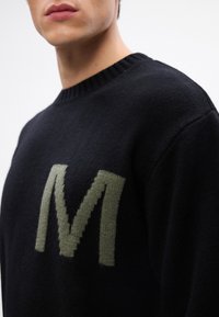 Suéter negro de punto con un gran logo verde de "M" en la parte frontal, con un cuello acanalado y una tela suave y texturizada.