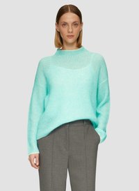 Pull en vert menthe léger, oversize, avec un col haut et une texture côtelée, associé à un pantalon gris sur mesure.