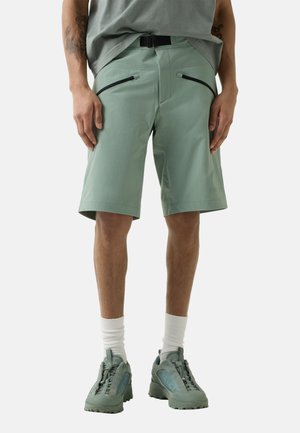 Man wearing light green knee-length shorts with black zipper pockets, white socks, and matching green hiking shoes on a plain background.