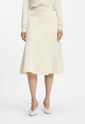 Kobieta ubrana w kremową spódnicę midi z bocznym paskiem, zestawioną z dopasowaną bluzką z długim rękawem oraz białymi butami na obcasie typu Mary Jane.