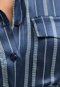 Close-up van een marineblauwe satijnen overhemd met witte touwachtige verticale strepen, donkerblauwe knopen en een knoopsluiting op de borstzak.