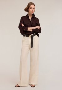 Camicia button-up marrone scuro con maniche arrotolate abbinata a pantaloni a gamba larga beige a vita alta e una cintura nera in contrasto.