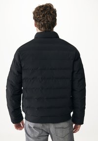 Muž s kudrnatými hnědými vlasy, oblečený v černé prošívané bundě a šedých džínách, viděn zezadu na bílém pozadí.