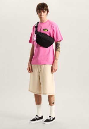 Sort skuldertaske med tekstureret overflade, båret over en lyserød skjorte. Cremefarvede shorts og sorte sneakers fuldender looket, med hvide sokker synlige.