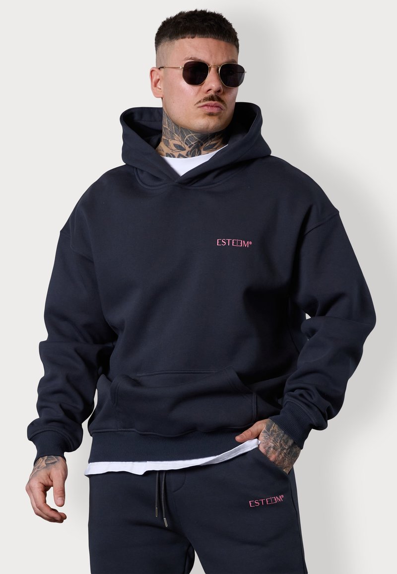 Sweatshirt à capuche bleu marine en tissu doux, doté d'une poche avant et d'un logo brodé rose. Porté avec un t-shirt blanc en dessous.